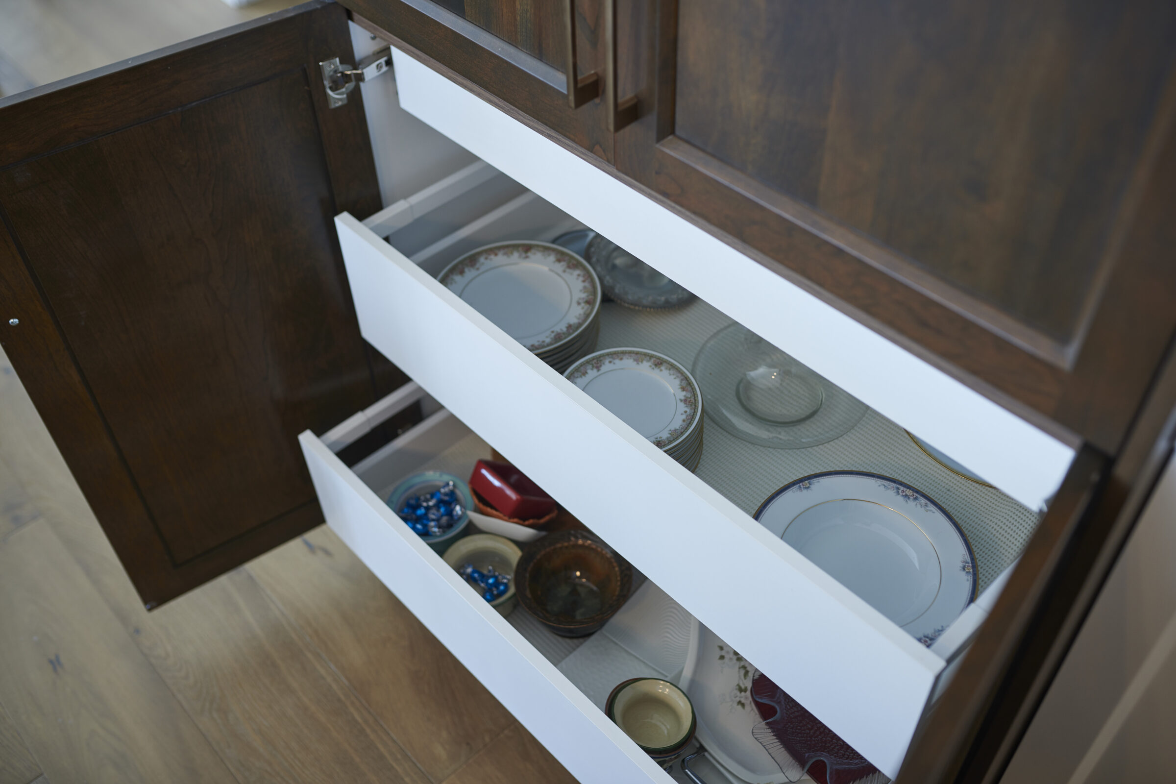 A kitchen cabinet with two drawers open, showcasing neatly organized dishes including plates, bowls, and a red container, against a wooden and white interior.