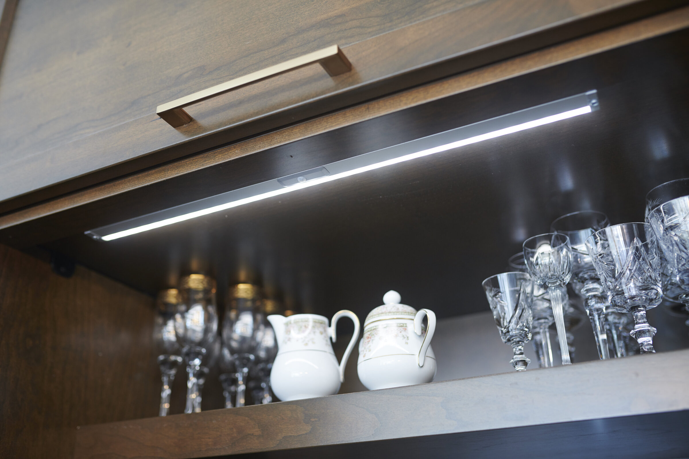 A dark wooden cabinet with an under-shelf light showcasing elegant glassware and a set of ornate white ceramic teapots. The ambiance is clean and sophisticated.