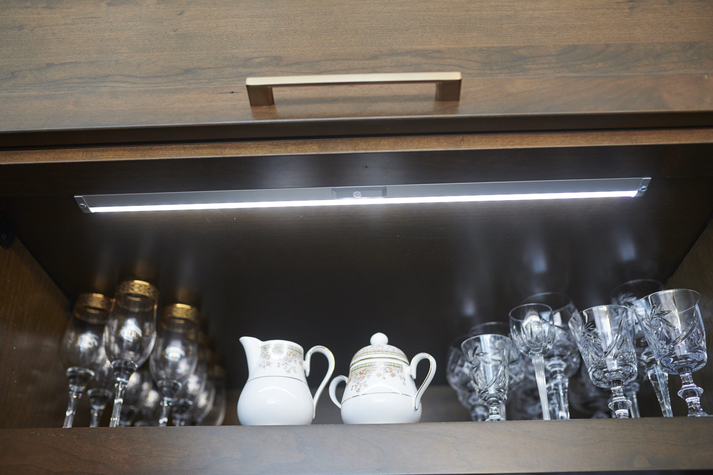 A wooden cabinet with an LED light illuminating glassware, including a teapot, cups, and ornate stemware, against a dark interior background.