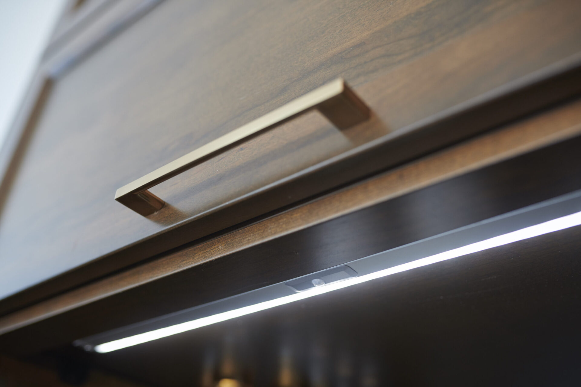 A close-up perspective of a wooden cabinet with a metallic handle, featuring under-cabinet lighting illuminating the countertop beneath with a soft glow.
