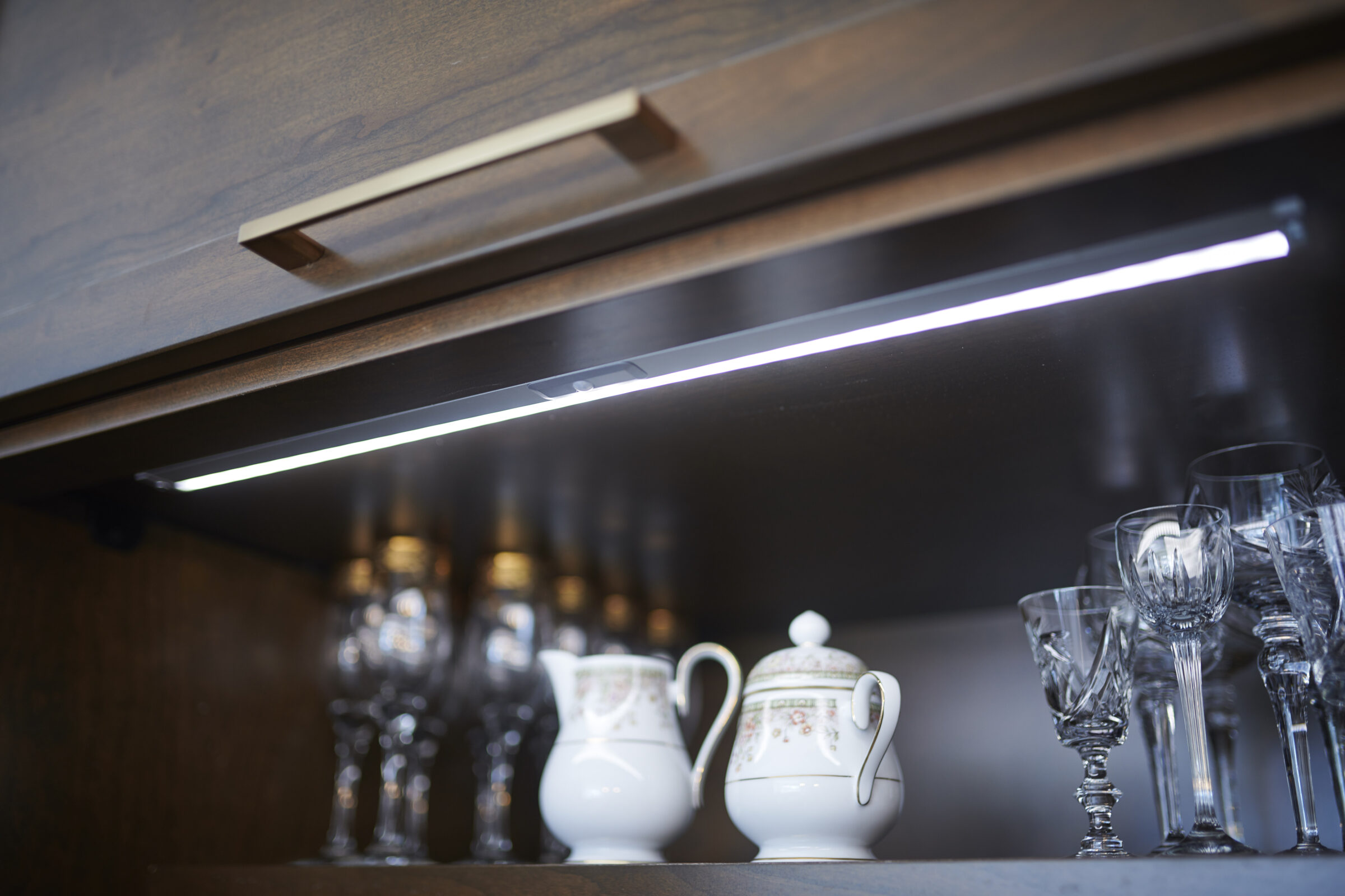 An under-cabinet LED light illuminates glassware and a porcelain teapot on a dark wood shelf, creating a bright, modern kitchen interior detail.