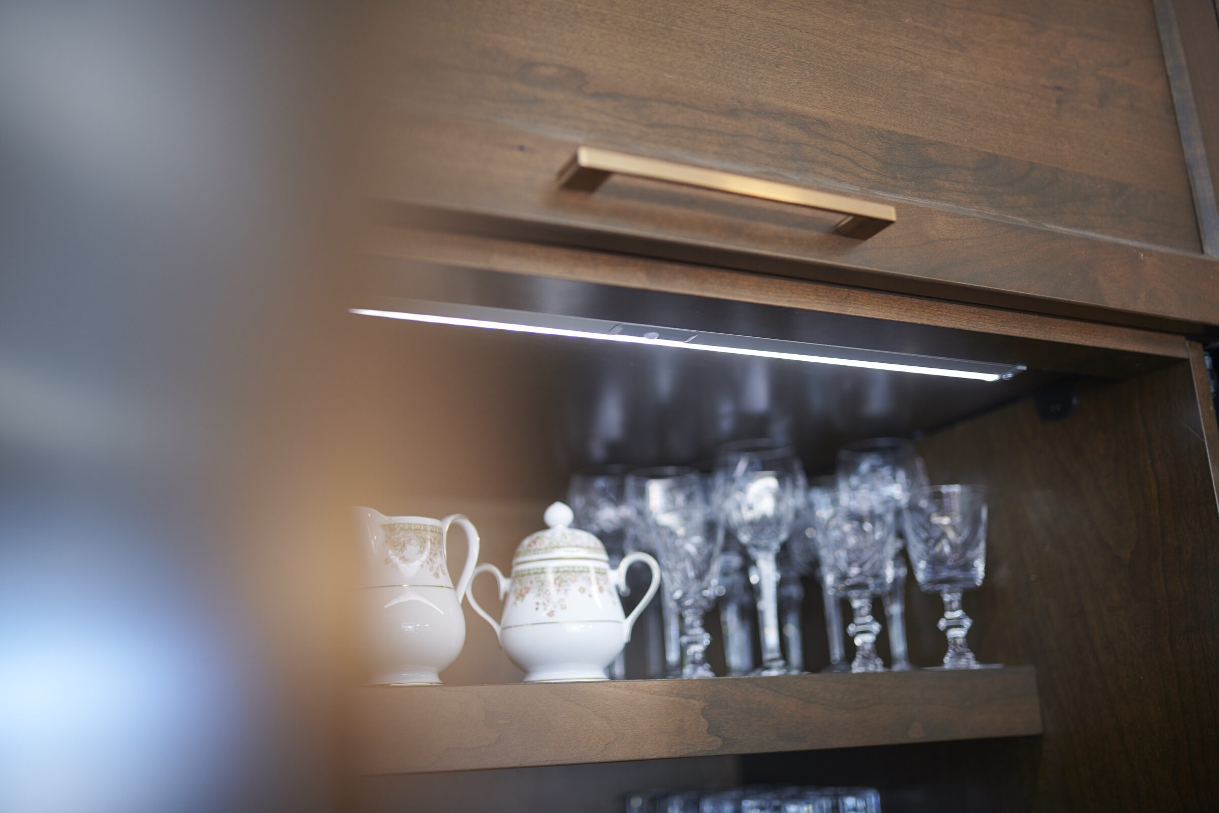 A wooden cabinet with an under-shelf light displaying a collection of glassware and a couple of decorative teapots, creating a warm, homey ambiance.