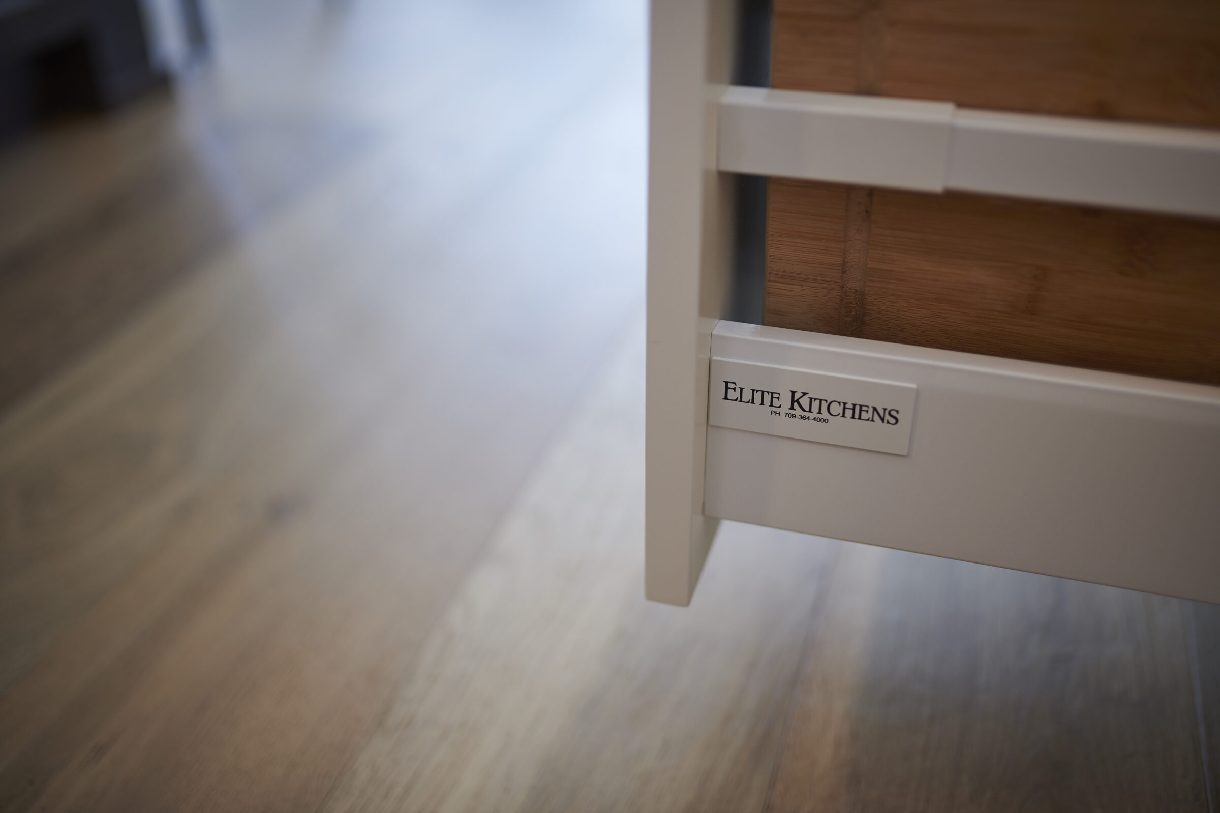 A close-up of a kitchen drawer with the text "Elite Kitchens" and a phone number. The background features a blurred wooden floor and cabinetry.