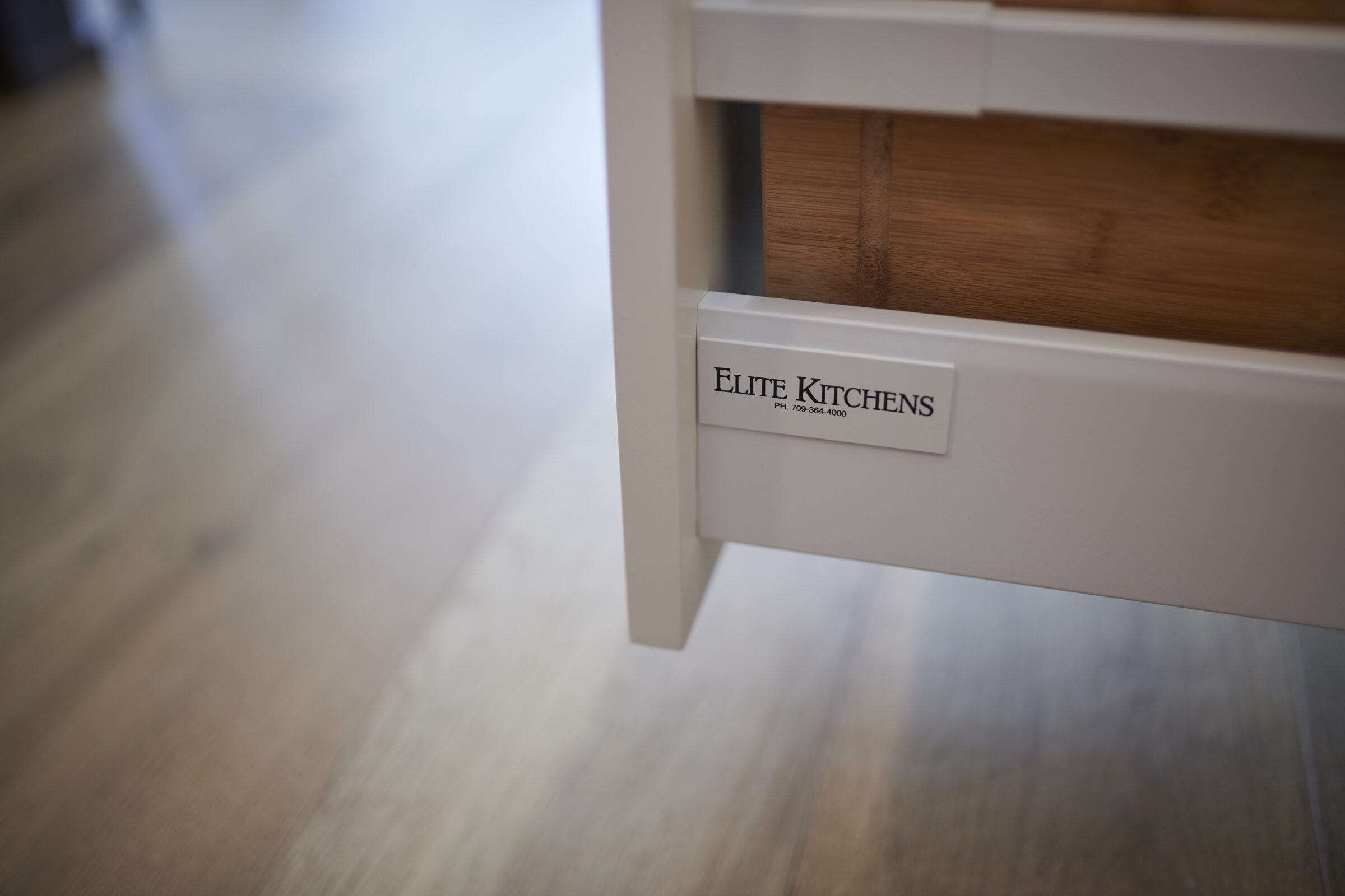 A close-up image of a white kitchen drawer with a label reading "Elite Kitchens" and a phone number, set against a wooden floor background.