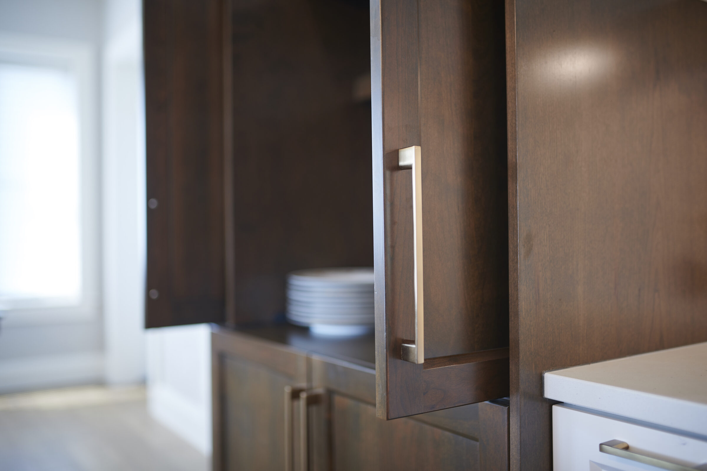 A modern kitchen with dark wood cabinets, a stack of white plates, a metal handle, and a blurred white countertop in the foreground.
