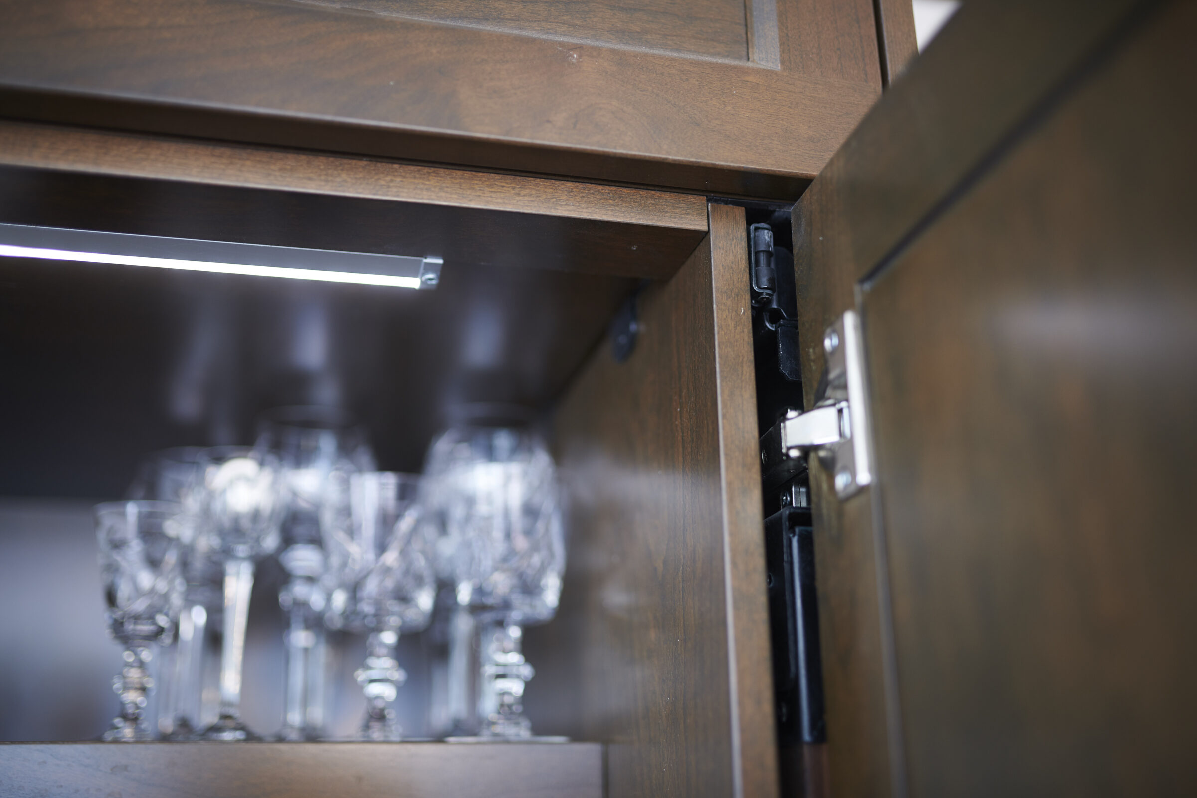 An open wooden cabinet with an under-cabinet light illuminating a collection of glassware inside, including what appear to be wine glasses.