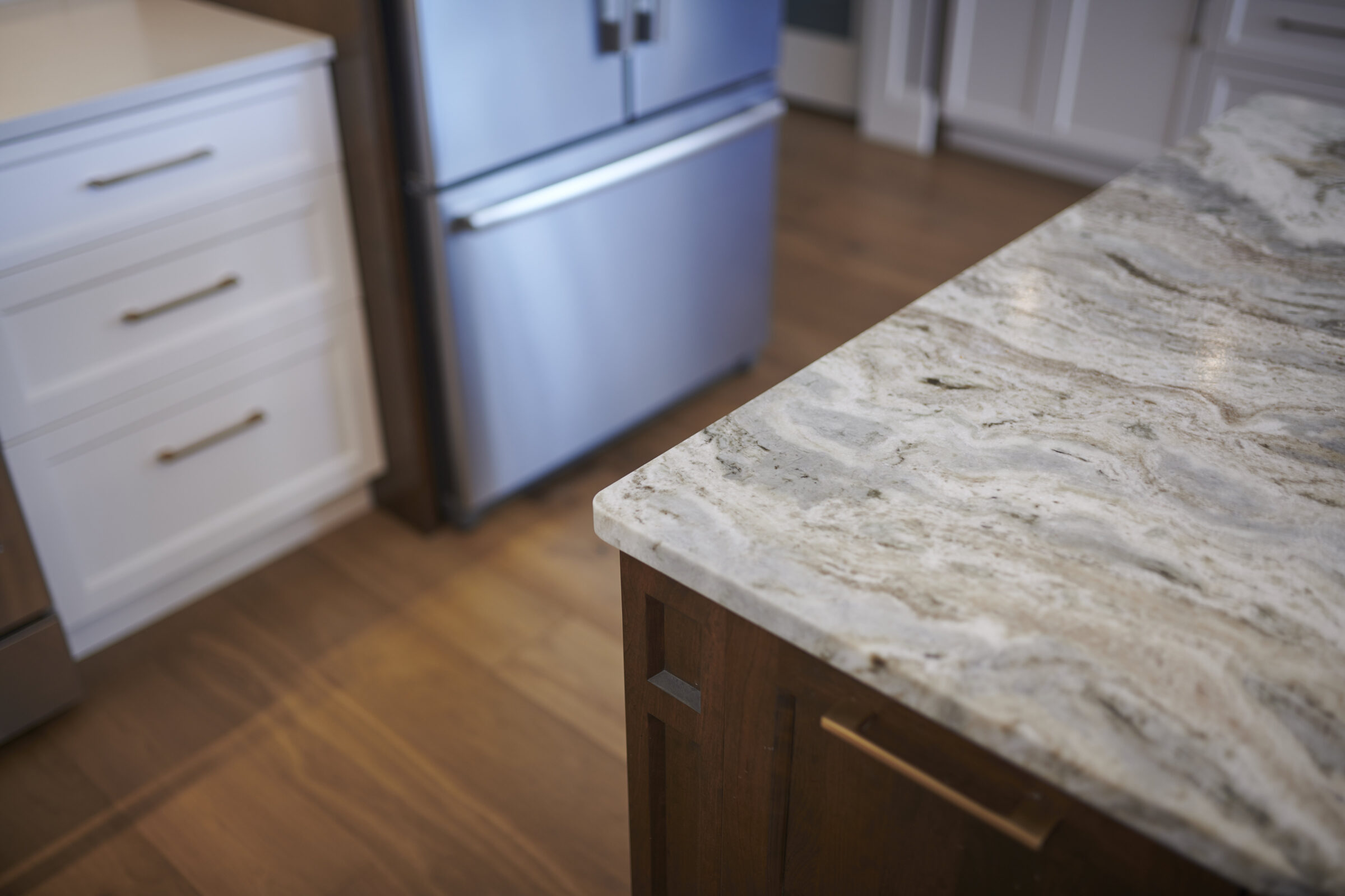 Modern kitchen interior with a close-up of a marble countertop extending over wooden cabinets. Stainless steel refrigerator and white drawers are visible in the background.