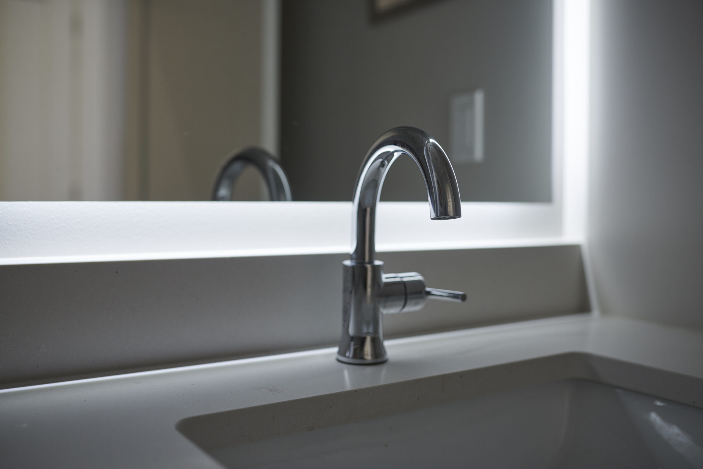A modern, sleek bathroom faucet with a silver finish stands atop a white sink countertop, reflecting in a mirror against a gray wall.