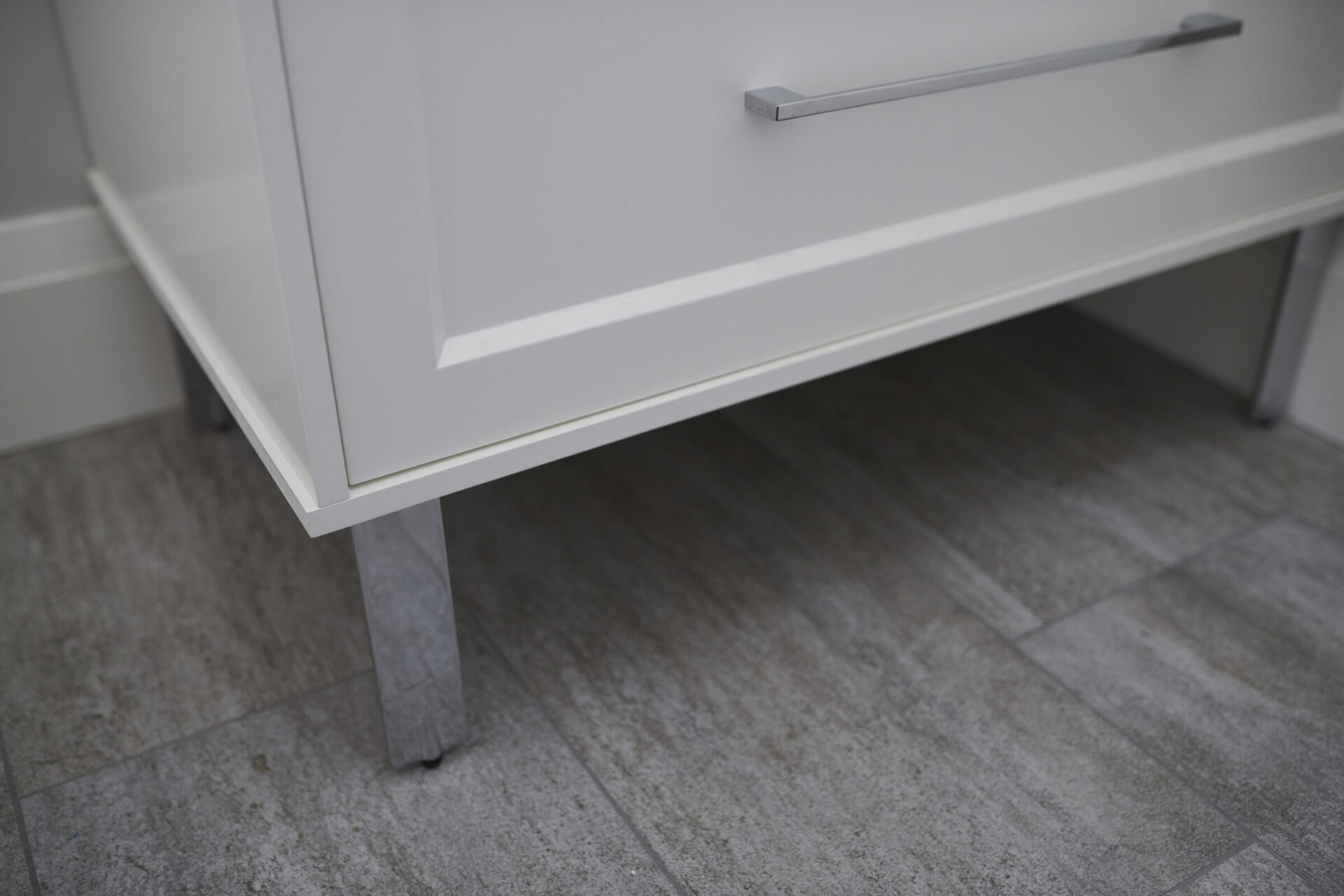A close-up view of a white cabinet with a modern handle, showing a partial drawer and leg, resting on a grey tiled floor.
