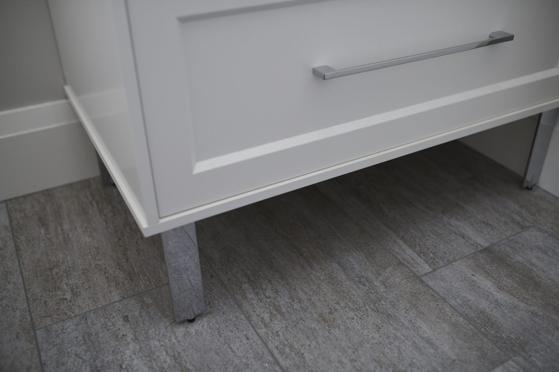 The image shows a close-up of a white cabinet with a metal handle over a gray tiled floor, focusing on the cabinet's base and leg.