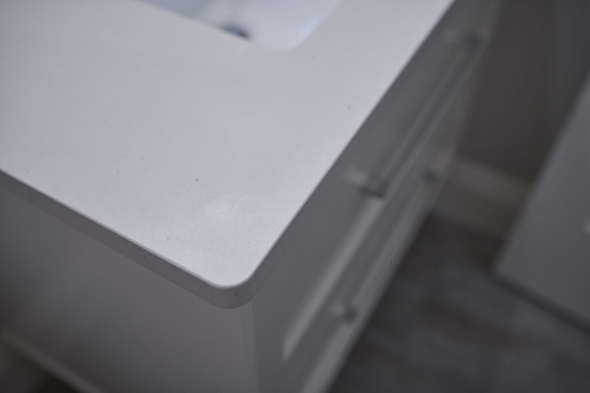 The image shows a close-up of a white countertop with a blurred background featuring grey cabinets, highlighting texture and subtle imperfections on the surface.