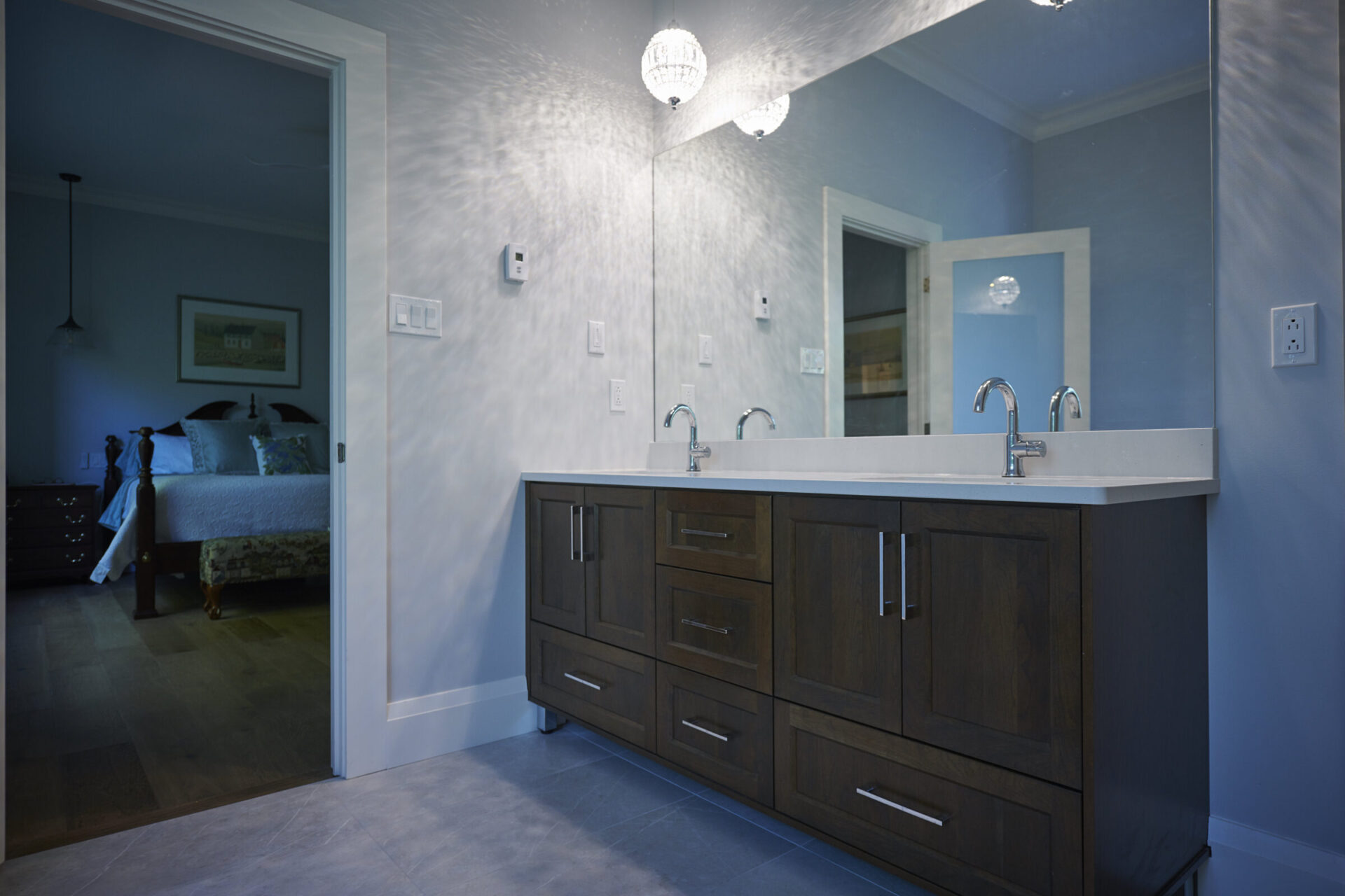 The image shows a modern bathroom with dark wood cabinets, a white countertop with two sinks, and a reflection of a bedroom through a mirror.