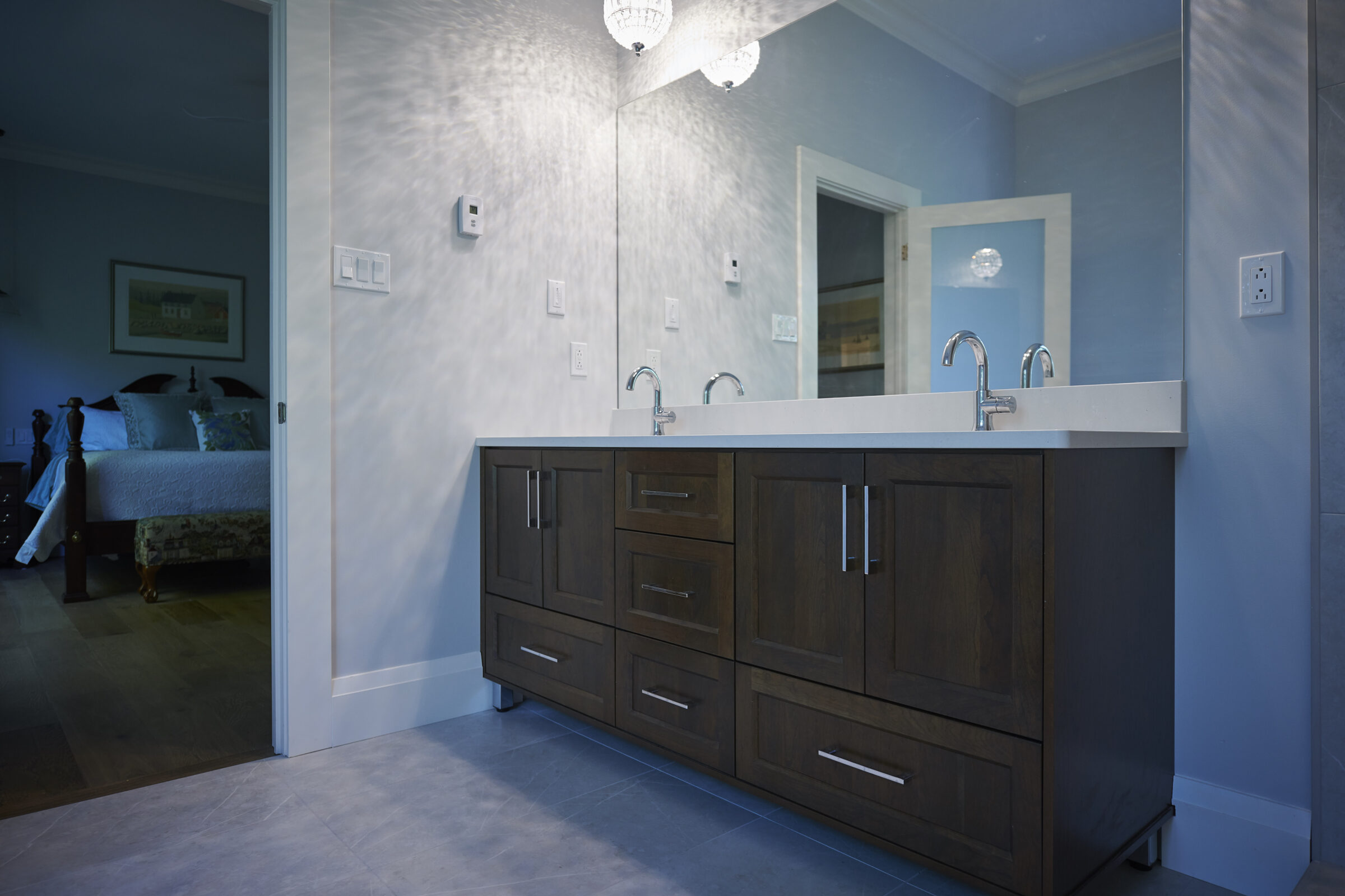 Modern bathroom with a dark wooden vanity, double sinks, and a chandelier. Light patterns on the wall. View into a bedroom through an open door.