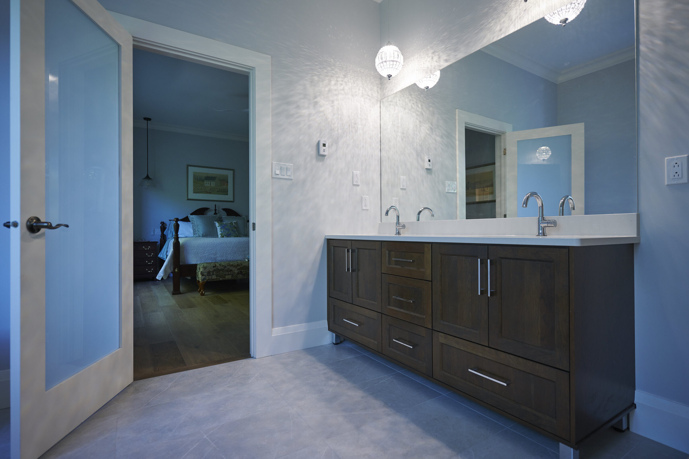 A modern bathroom with a dark wooden vanity, dual sinks, and a large mirror. The room leads to a bedroom with a visible bed.