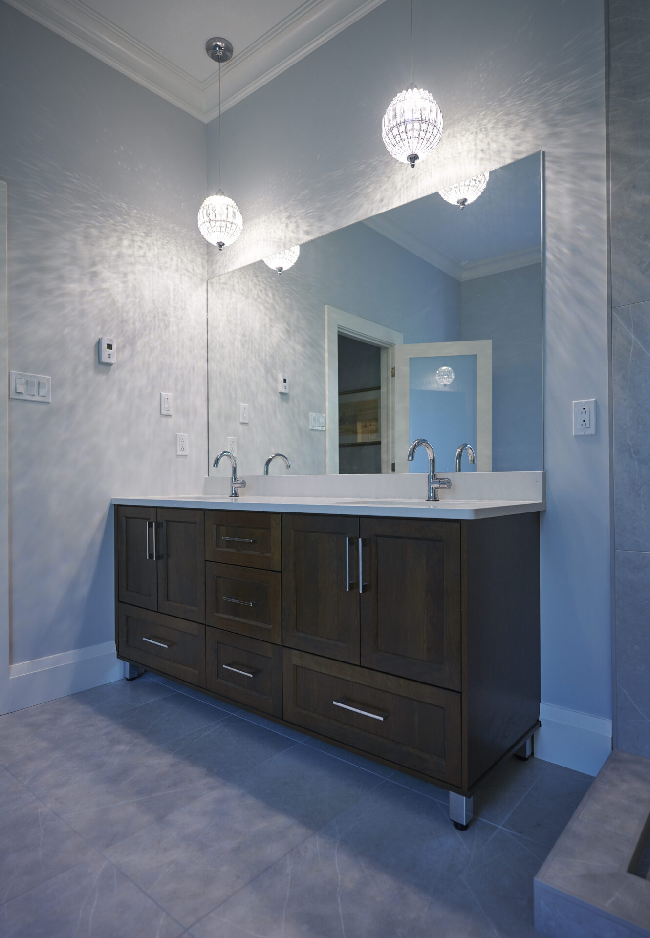 A modern bathroom with a dark wood double vanity, white countertop, two sinks, wall-mounted faucets, and ornate pendant lights casting a patterned glow.