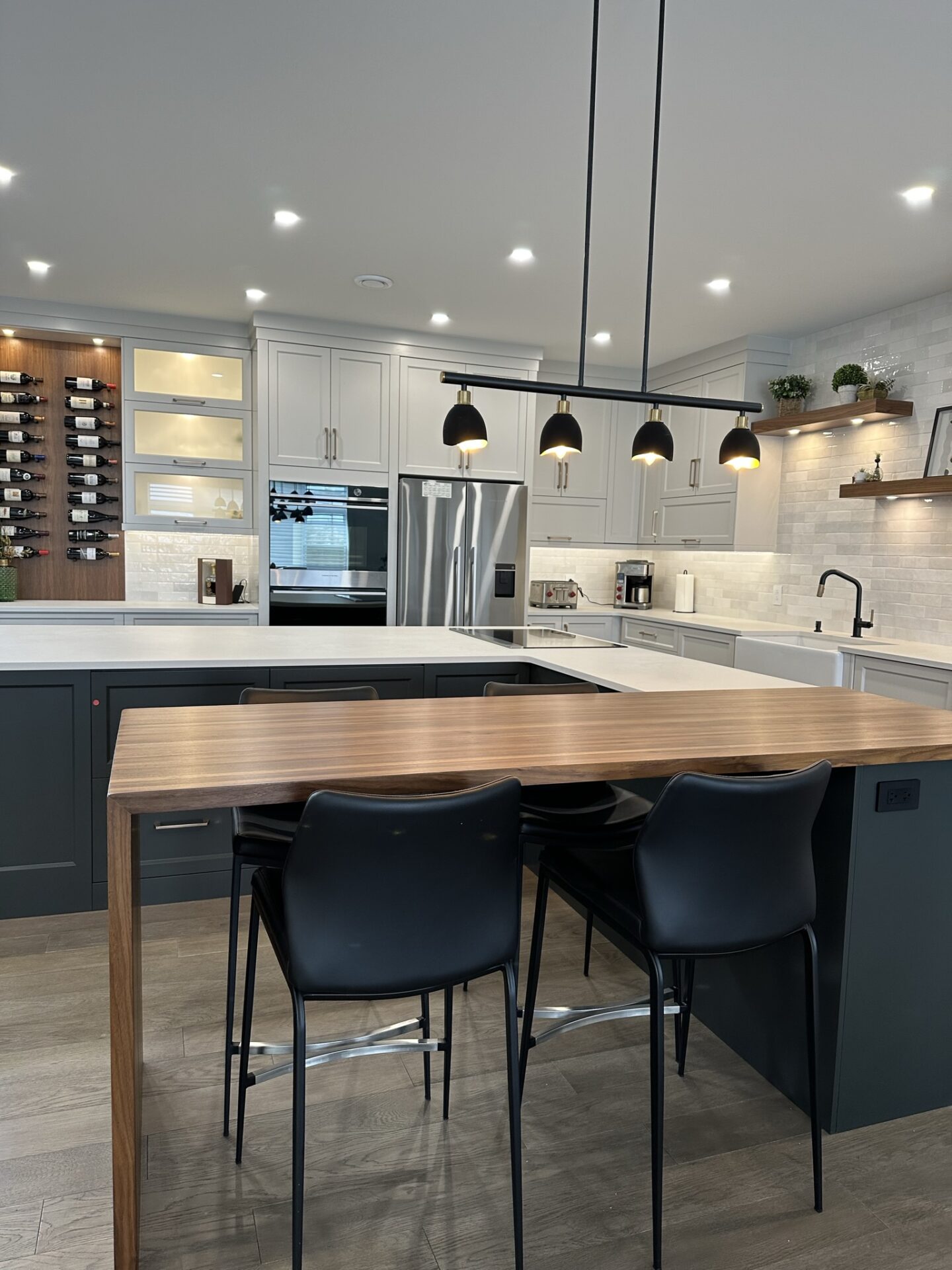 Modern kitchen featuring a large center island with a wood dining extension, matte black chairs, sleek black pendant lights, and a wine display wall with white cabinetry, stainless steel appliances, and open wood shelving.