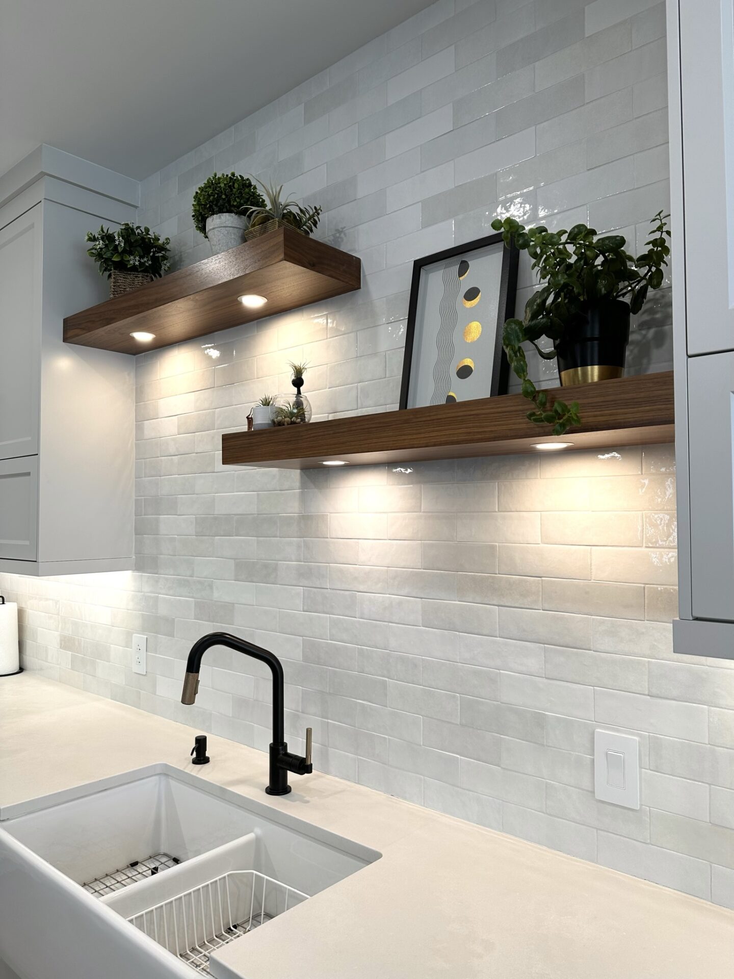 Contemporary kitchen backsplash with floating wood shelves and under-cabinet lighting. Decor includes potted plants, framed art, and a matte black faucet over a white apron sink. The glossy subway tile adds a clean, modern look.