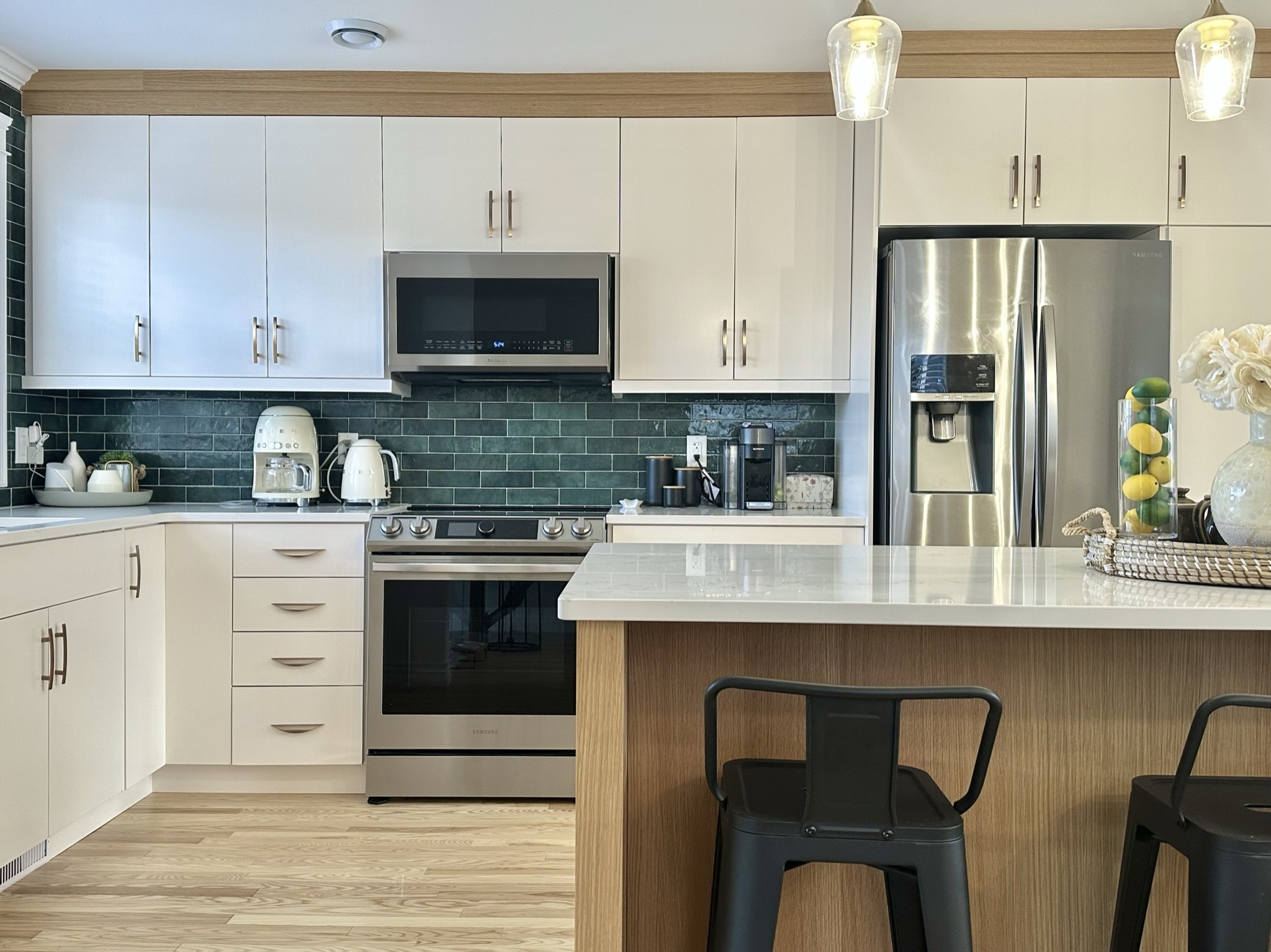 Contemporary kitchen with glossy white cabinets, stainless steel appliances, wood accents, and a dark green subway tile backsplash, featuring a white quartz island with black stools.
