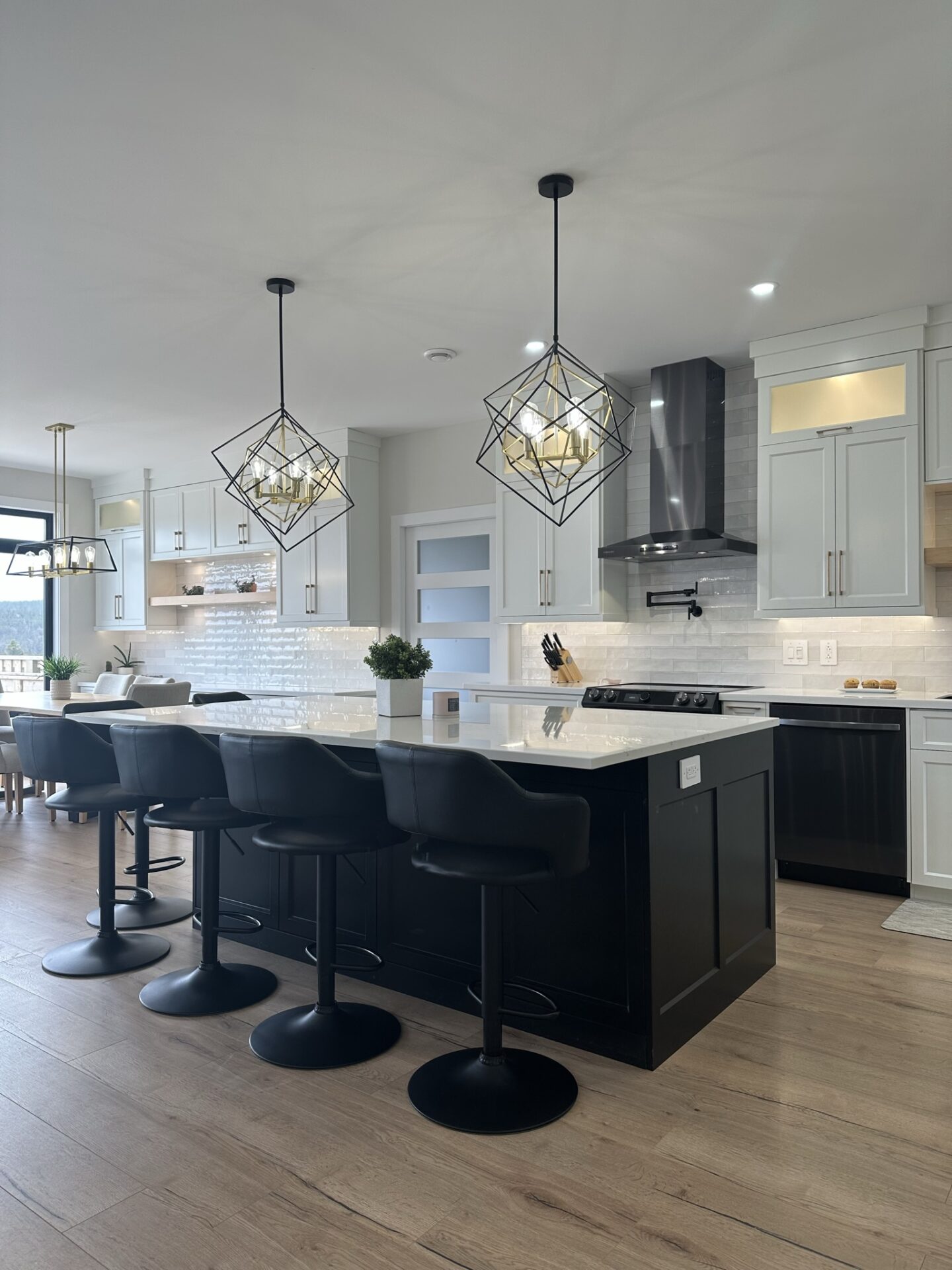 Modern kitchen interior with a large island featuring black cabinetry and four black barstools, pendant lighting with geometric metal frames, white upper cabinets, light wood flooring, and stainless steel appliances in a bright, open layout.