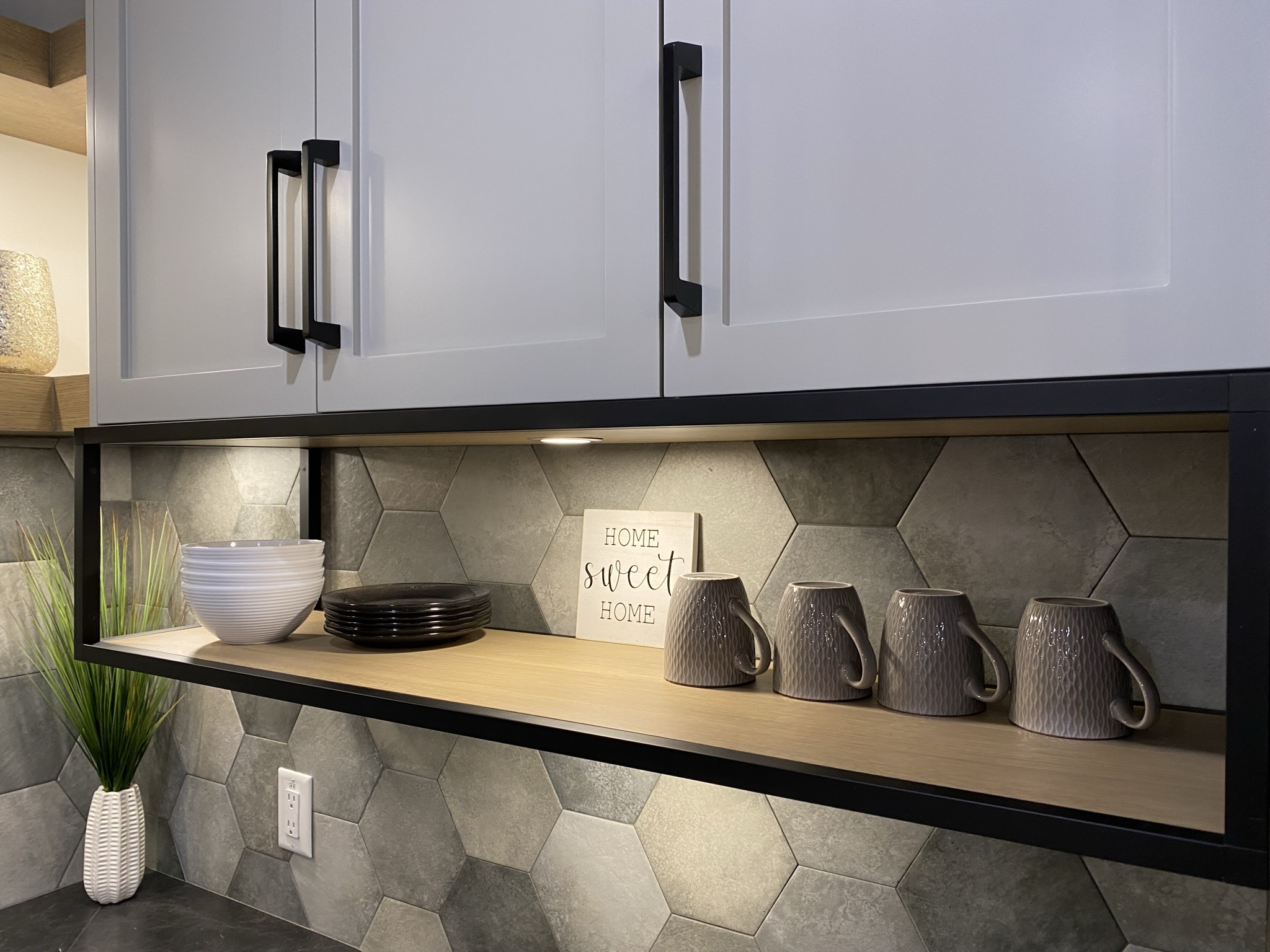 Modern kitchen shelf display with stacked white bowls, black plates, taupe ceramic mugs, a small "Home Sweet Home" sign, and under-cabinet lighting against a hexagonal tile backsplash.