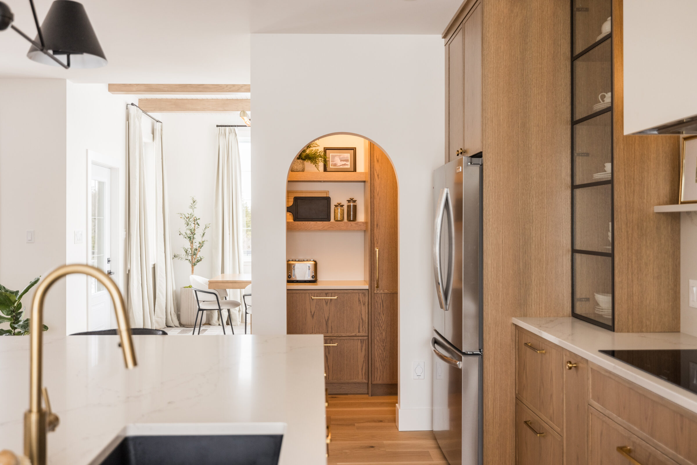 Warm, modern kitchen with natural wood cabinetry, a gold faucet over a marble island, and a built-in arched pantry nook featuring open shelves with small appliances and decorative accents.