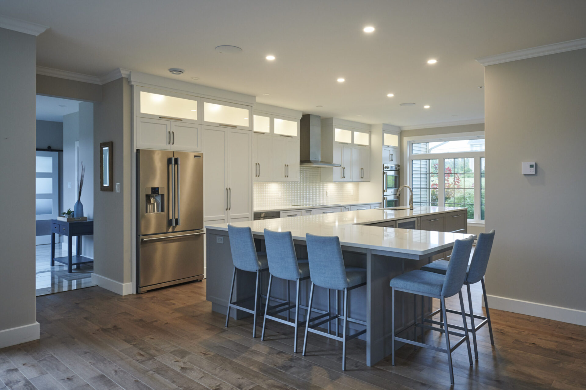 Spacious transitional kitchen with white cabinetry, a large island featuring light blue upholstered bar stools, and stainless steel appliances. Recessed lighting and glass-front upper cabinets brighten the space, while wide-plank wood flooring adds warmth. A wall of windows brings in natural light from the backyard.