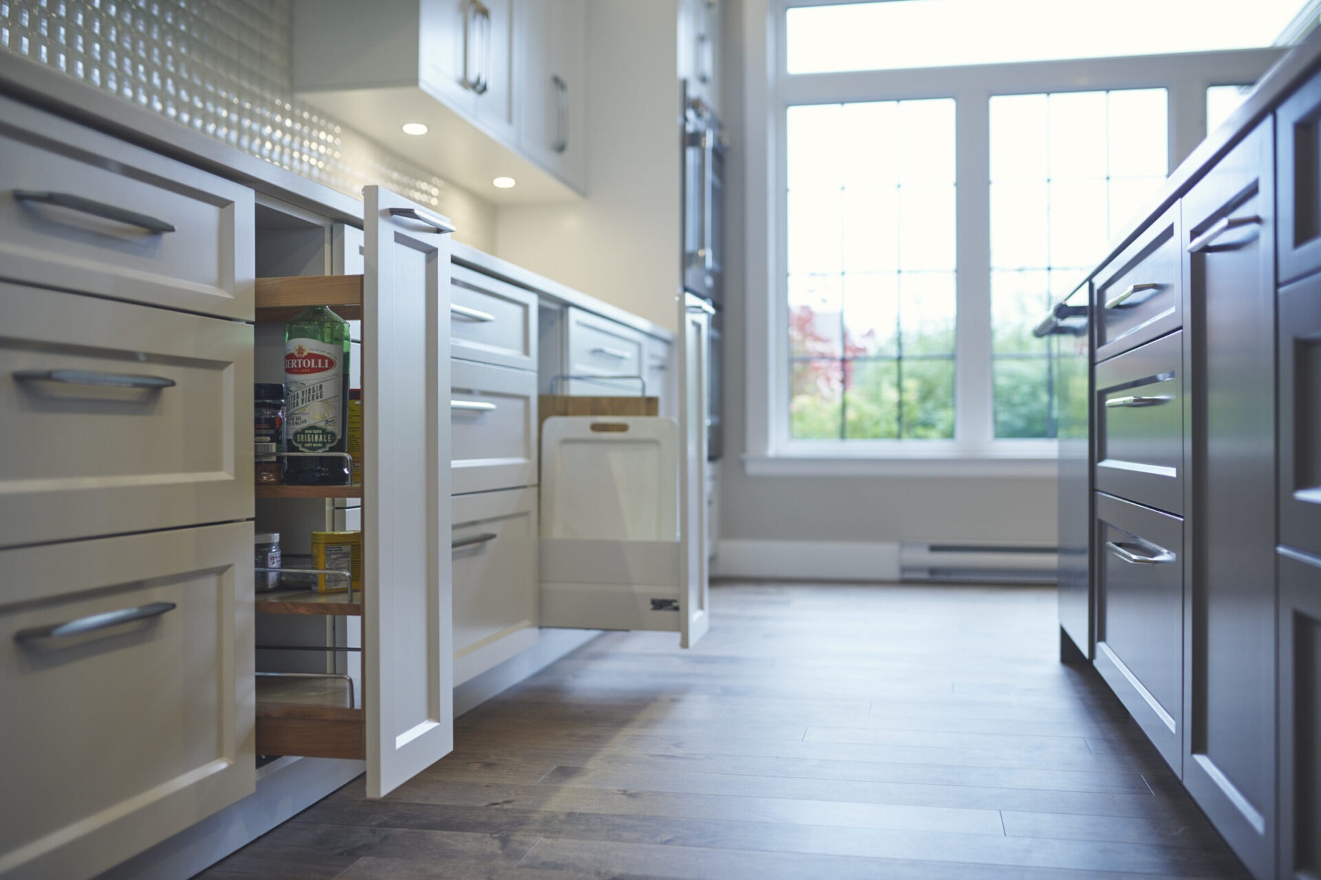 Modern kitchen with white cabinets and hardwood floors, featuring pull-out pantry and waste bin drawers, with natural light streaming through large windows.