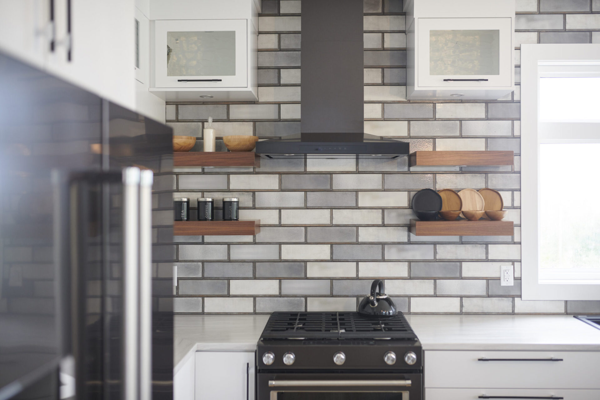 Contemporary kitchen with glossy gray subway tile backsplash, black range hood, matte black stove, and floating wood shelves holding spice jars, bowls, and decor items.
