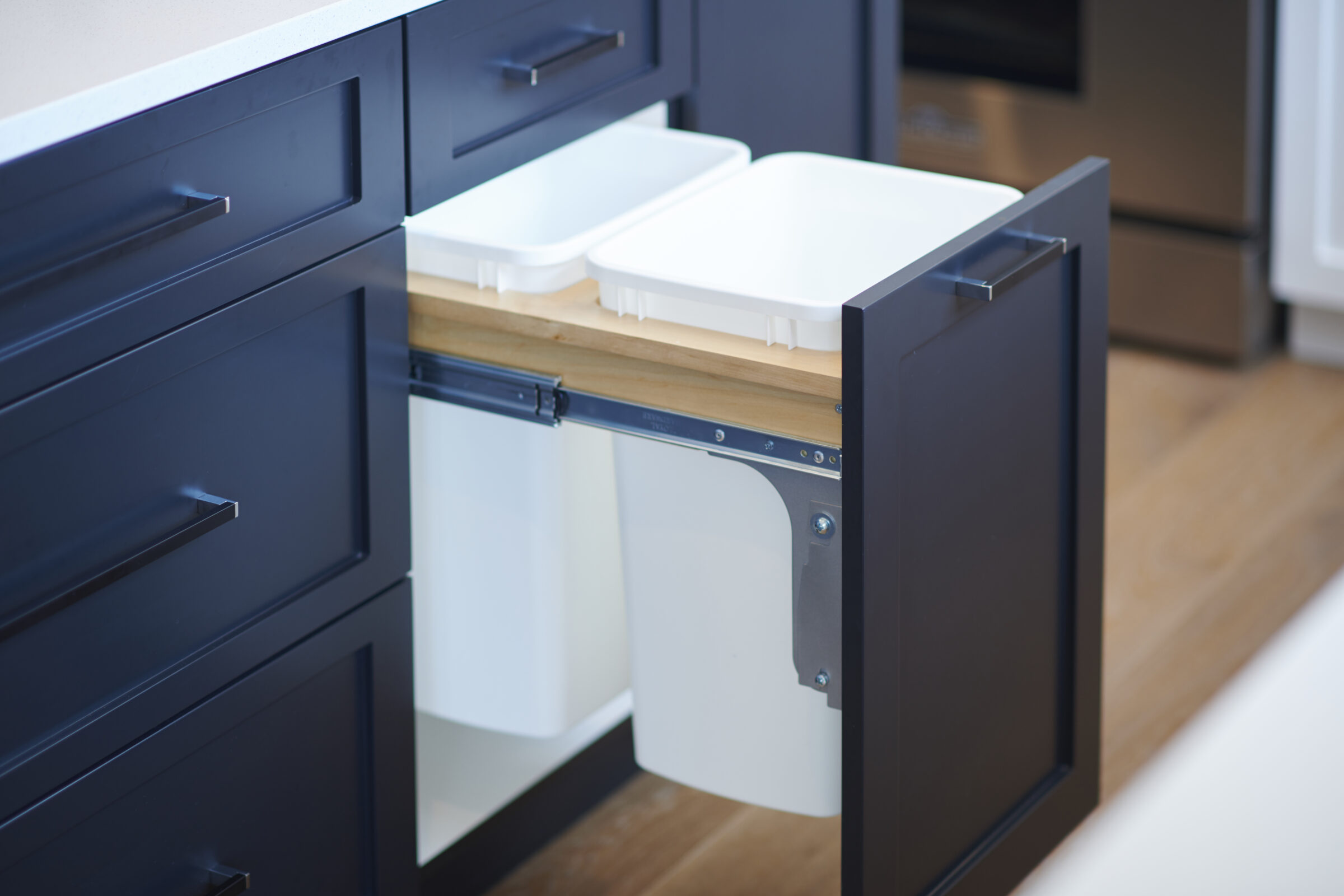 Pull-out kitchen cabinet with dark blue paneling revealing dual white bins for waste and recycling, neatly built into the drawer for hidden trash storage.