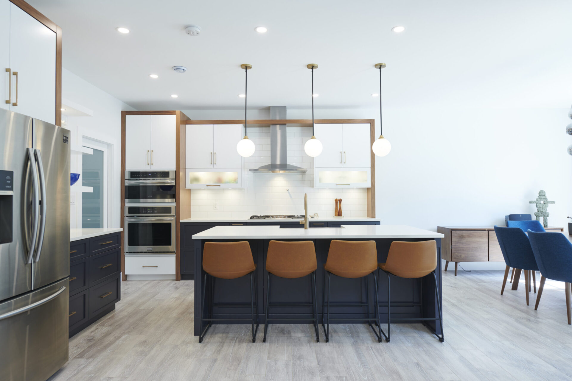 Contemporary kitchen with a bright, minimalist design featuring a central island with brown bar stools, globe pendant lighting, white upper cabinets, navy blue lower cabinets, and stainless steel appliances. The open-concept layout includes a dining area with blue chairs and wood accents.