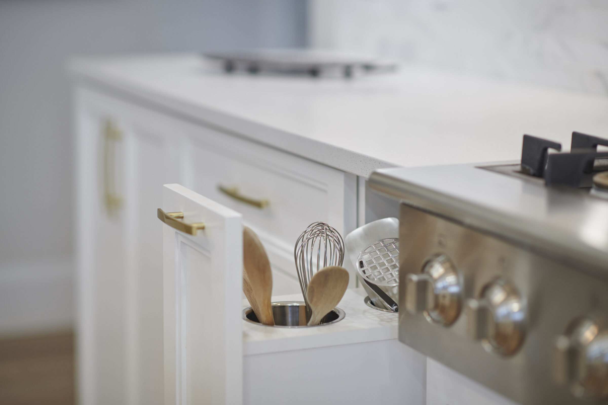 Pull-out kitchen cabinet organizer next to a stainless steel stove, featuring built-in canisters holding wooden spoons, a whisk, and metal cooking utensils for easy access.