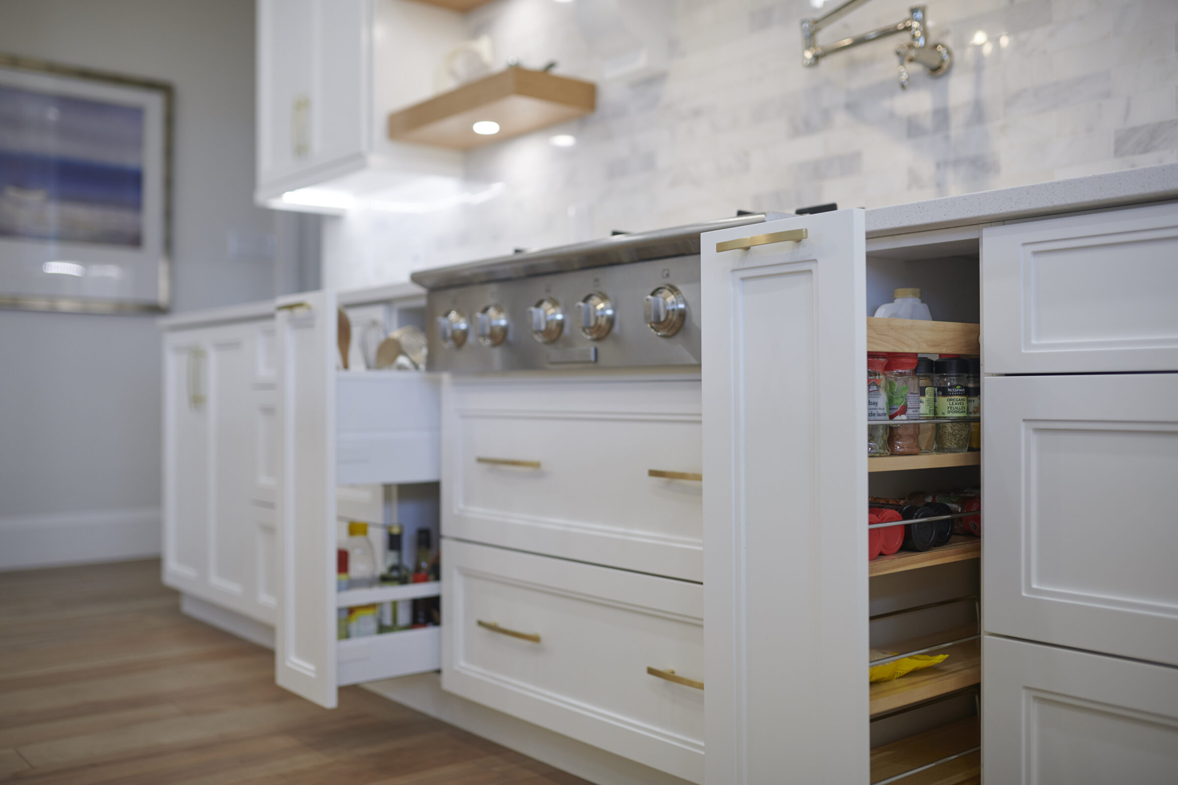 Close-up of a modern white kitchen with pull-out pantry drawers open on either side of a stainless steel stove, revealing neatly organized spices and cooking oils.