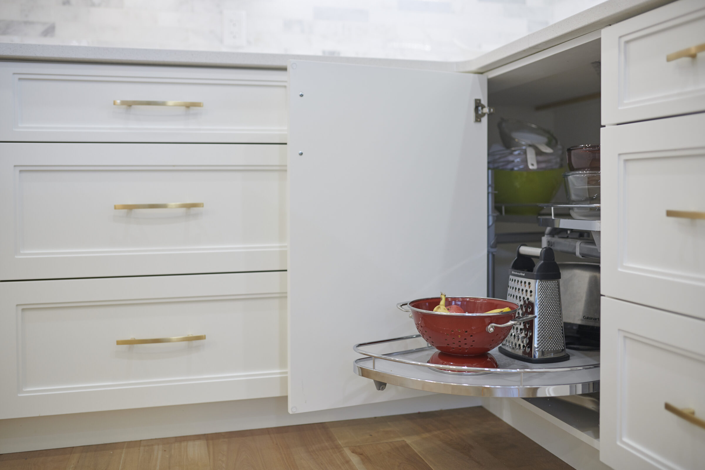 White kitchen base cabinet with a swing-out corner shelf extended, displaying a red colander with fruit, a box grater, and organized storage of glassware and appliances inside.