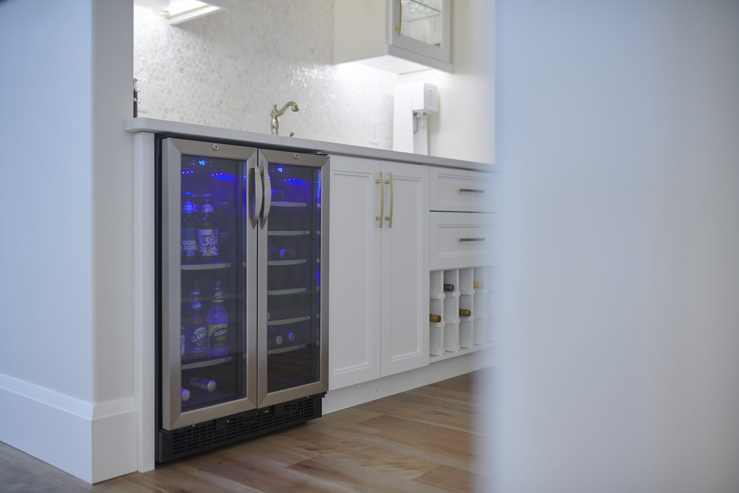 Modern home bar area with a dual-zone beverage cooler lit by blue LED lights, white cabinetry with gold hardware, a wine rack, and a small sink under a mosaic tile backsplash.