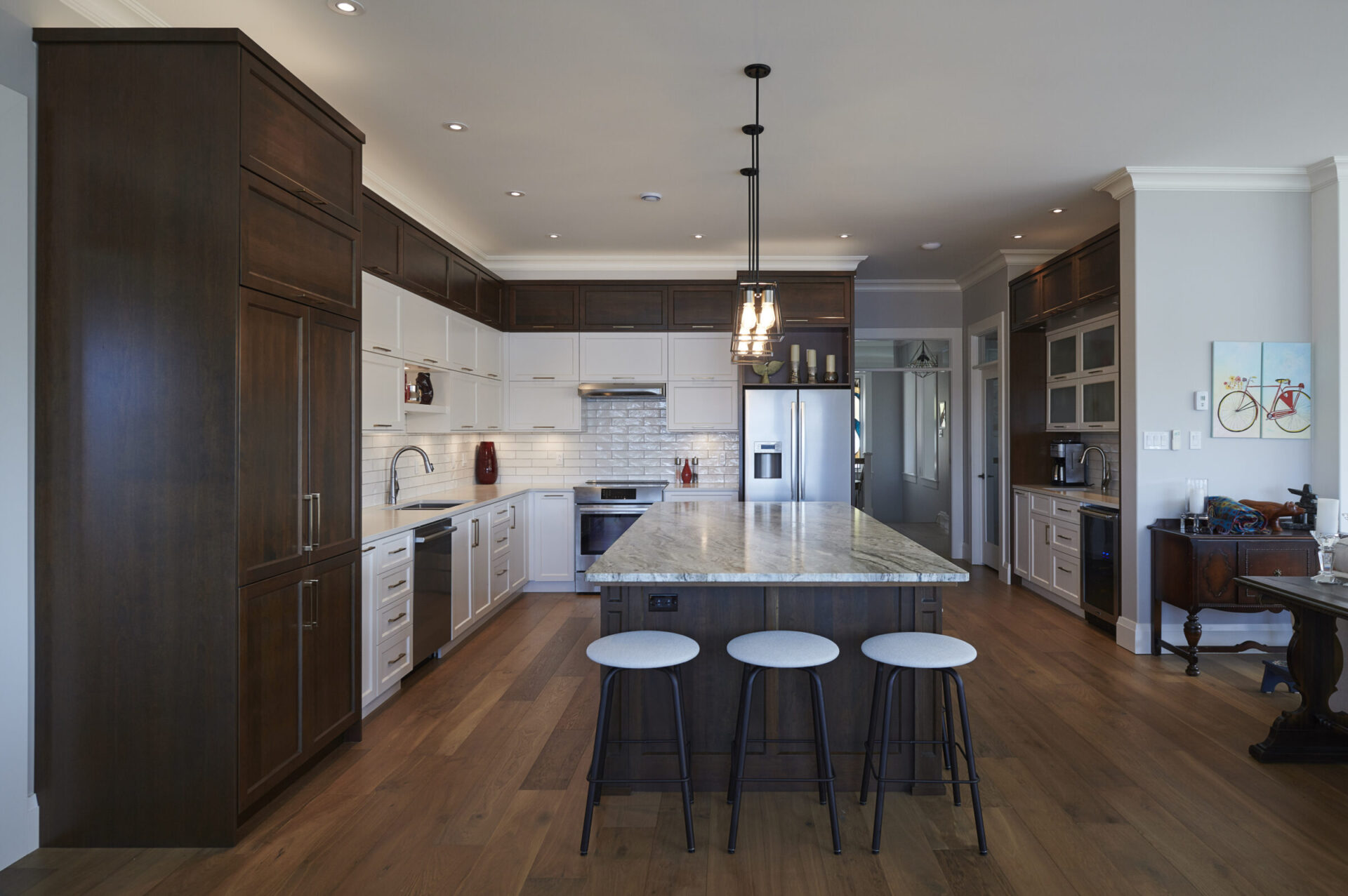 Spacious transitional kitchen featuring a large island with a granite countertop and three modern barstools. The space combines dark wood cabinetry with white shaker cabinets, stainless steel appliances, subway tile backsplash, and warm hardwood flooring. Pendant lighting adds a touch of elegance above the island.