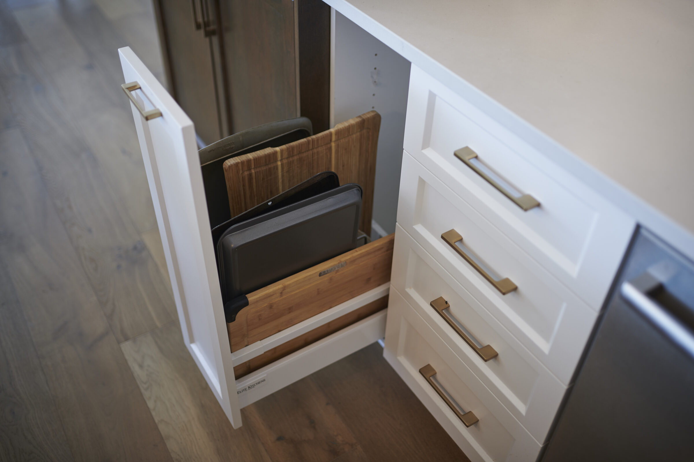 Pull-out cabinet organizer in a modern kitchen featuring vertical slots for neatly storing cutting boards, baking sheets, and trays, with white cabinetry and gold hardware.