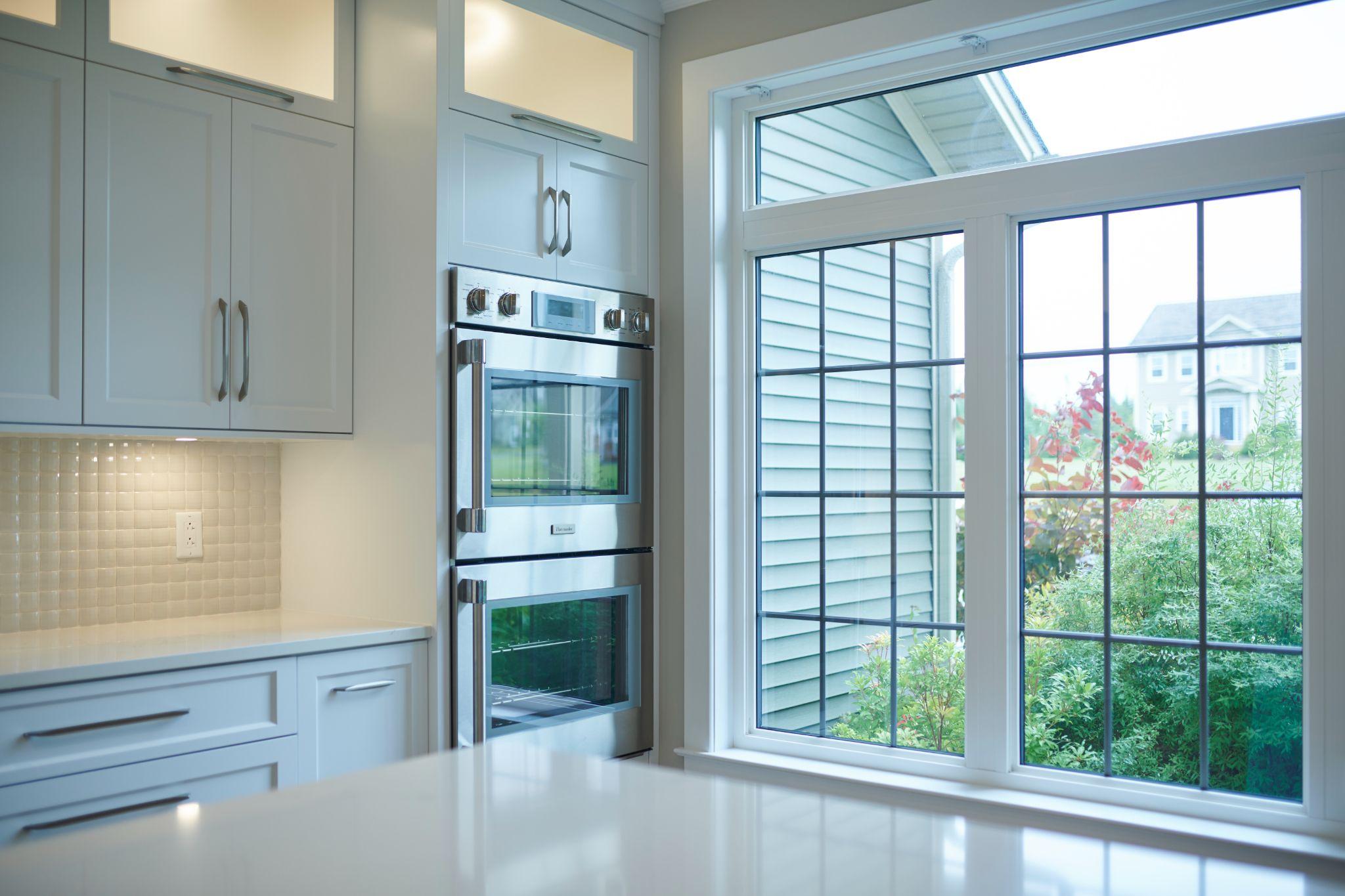 Bright kitchen corner with white cabinetry, mosaic tile backsplash, and built-in stainless steel double ovens positioned beside a large window overlooking a green yard and neighboring house.