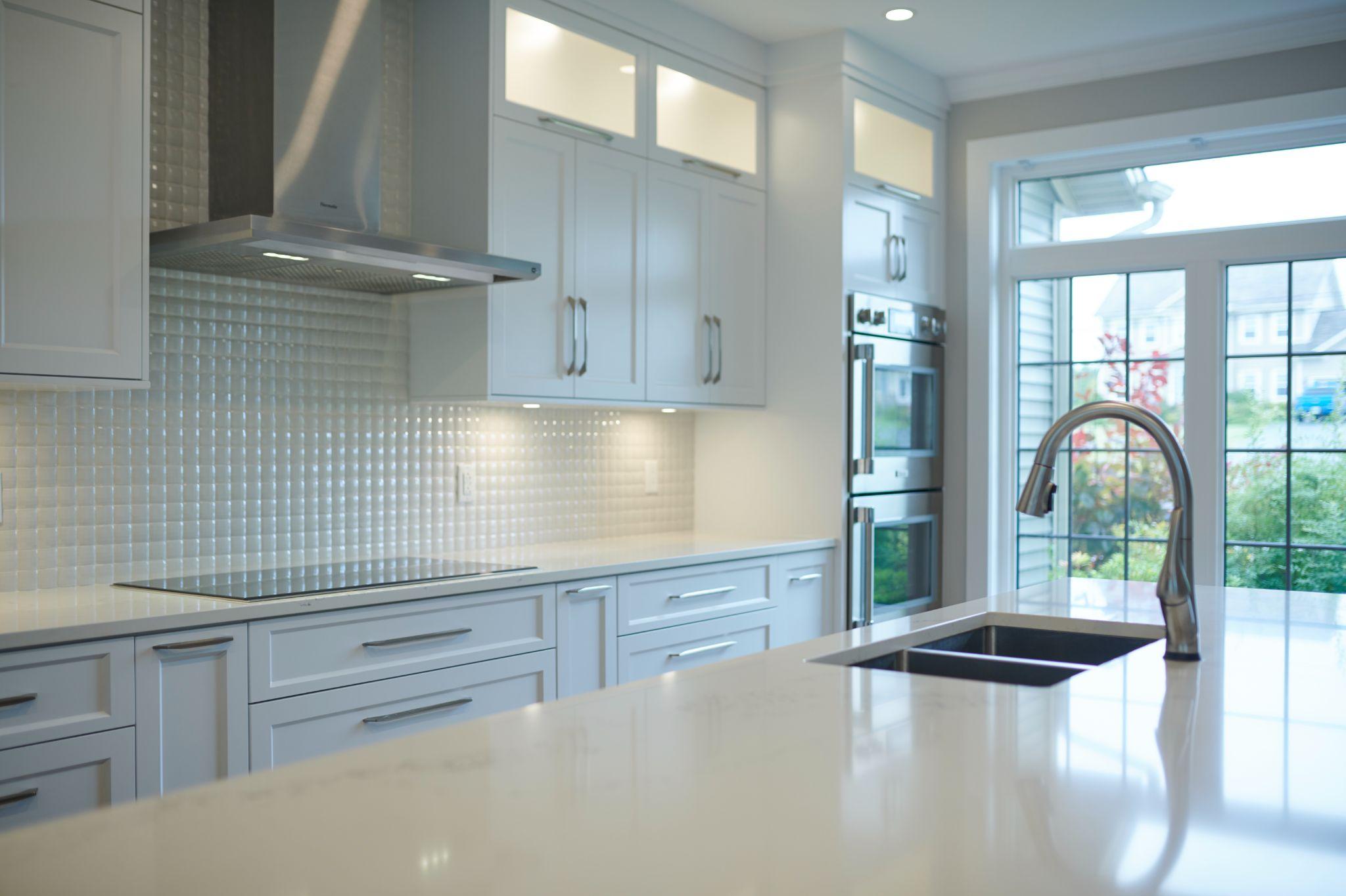 Bright modern kitchen with glossy white cabinetry, stainless steel appliances, mosaic tile backsplash, a sleek induction cooktop, and a large island with built-in sink beneath tall windows.