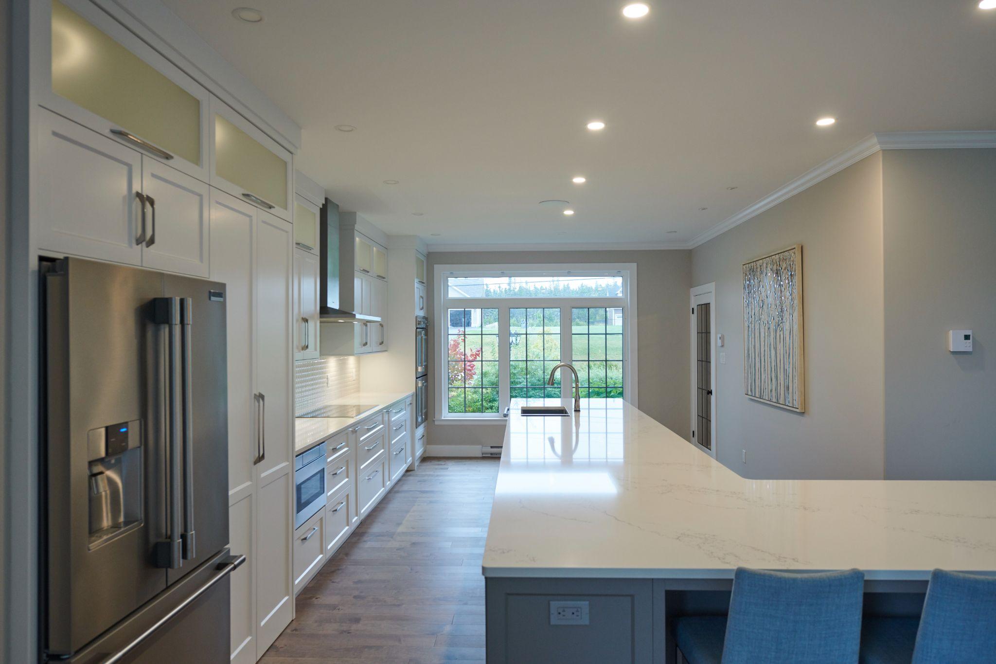 Long, modern kitchen with large quartz island, white cabinets, stainless steel appliances, and a wall of windows overlooking a green backyard.