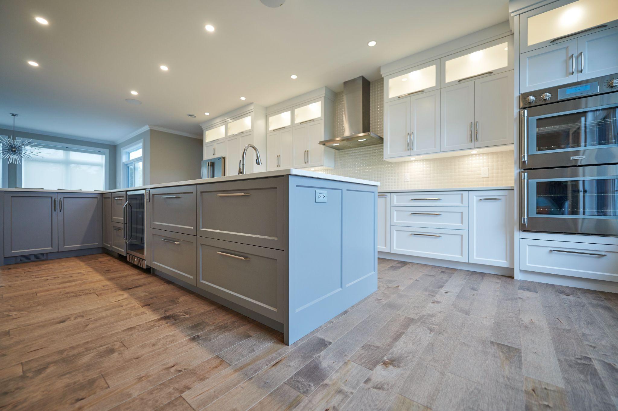 Low-angle view of a large kitchen island with gray cabinetry, quartz countertop, and built-in wine cooler, surrounded by white cabinets and wood flooring.