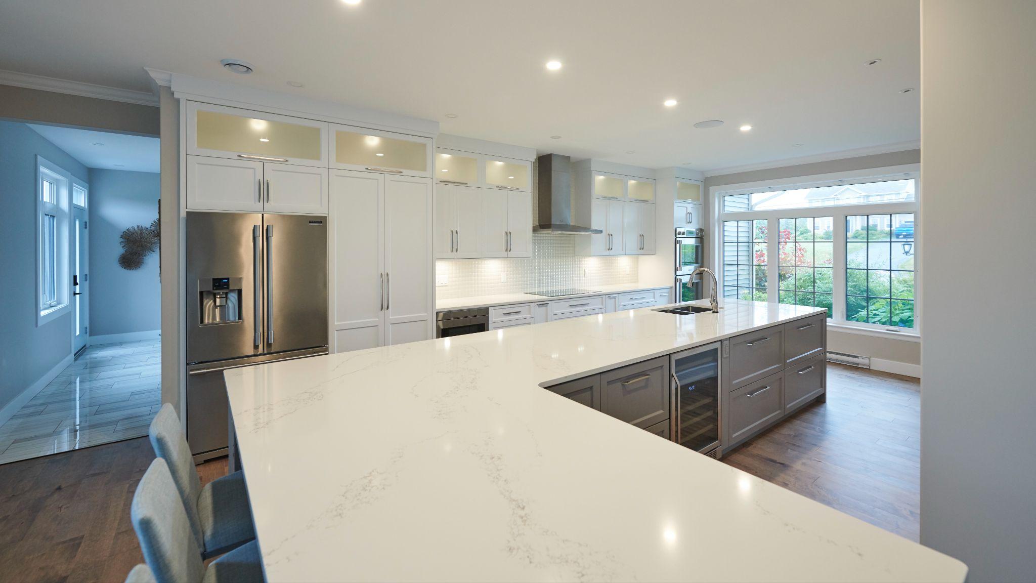 Modern kitchen with a large U-shaped white quartz island, stainless steel fridge, white cabinetry, and a wall of windows providing natural light.