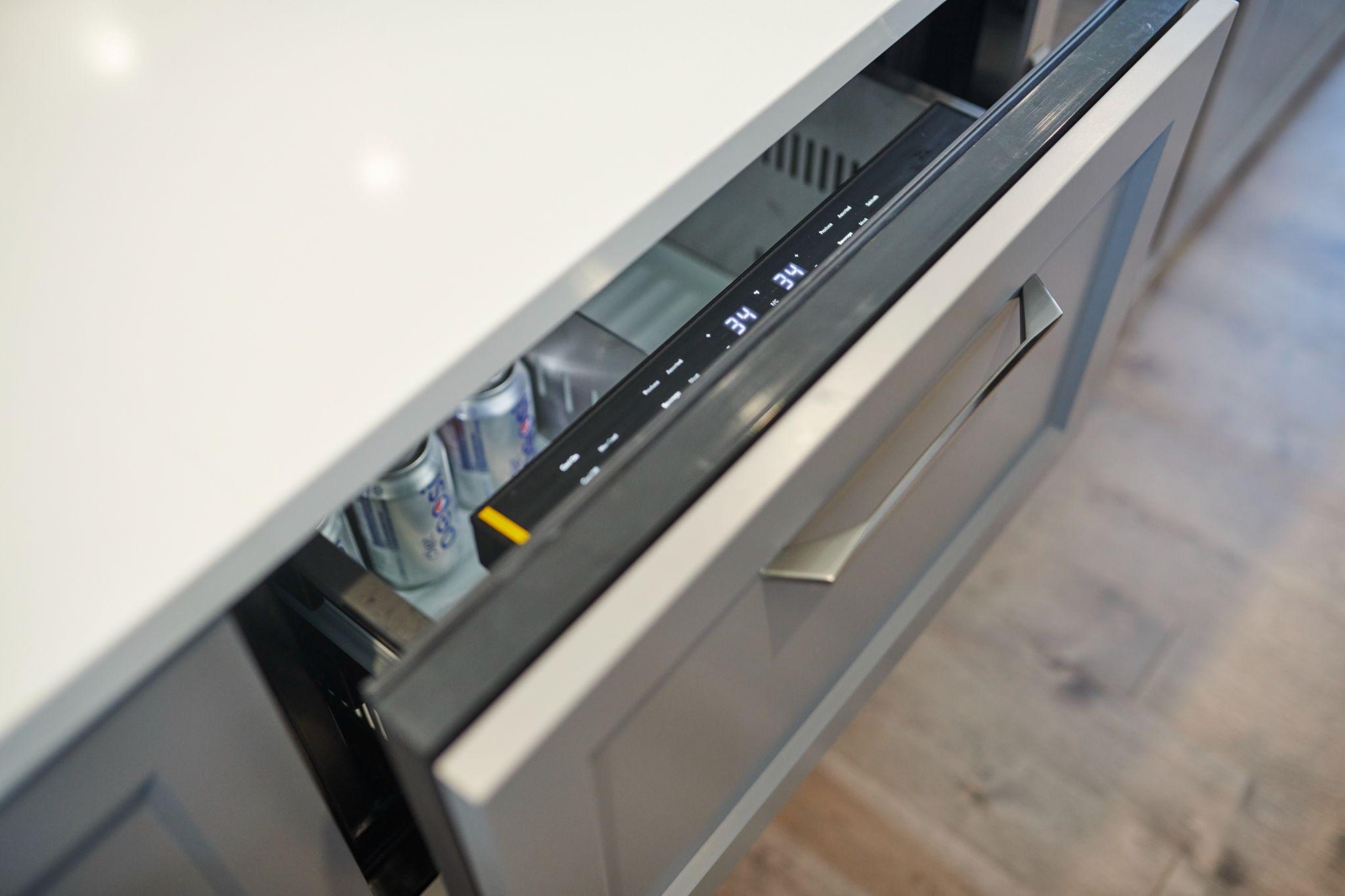 Open drawer-style beverage fridge built into a gray kitchen island, partially filled with soda cans.