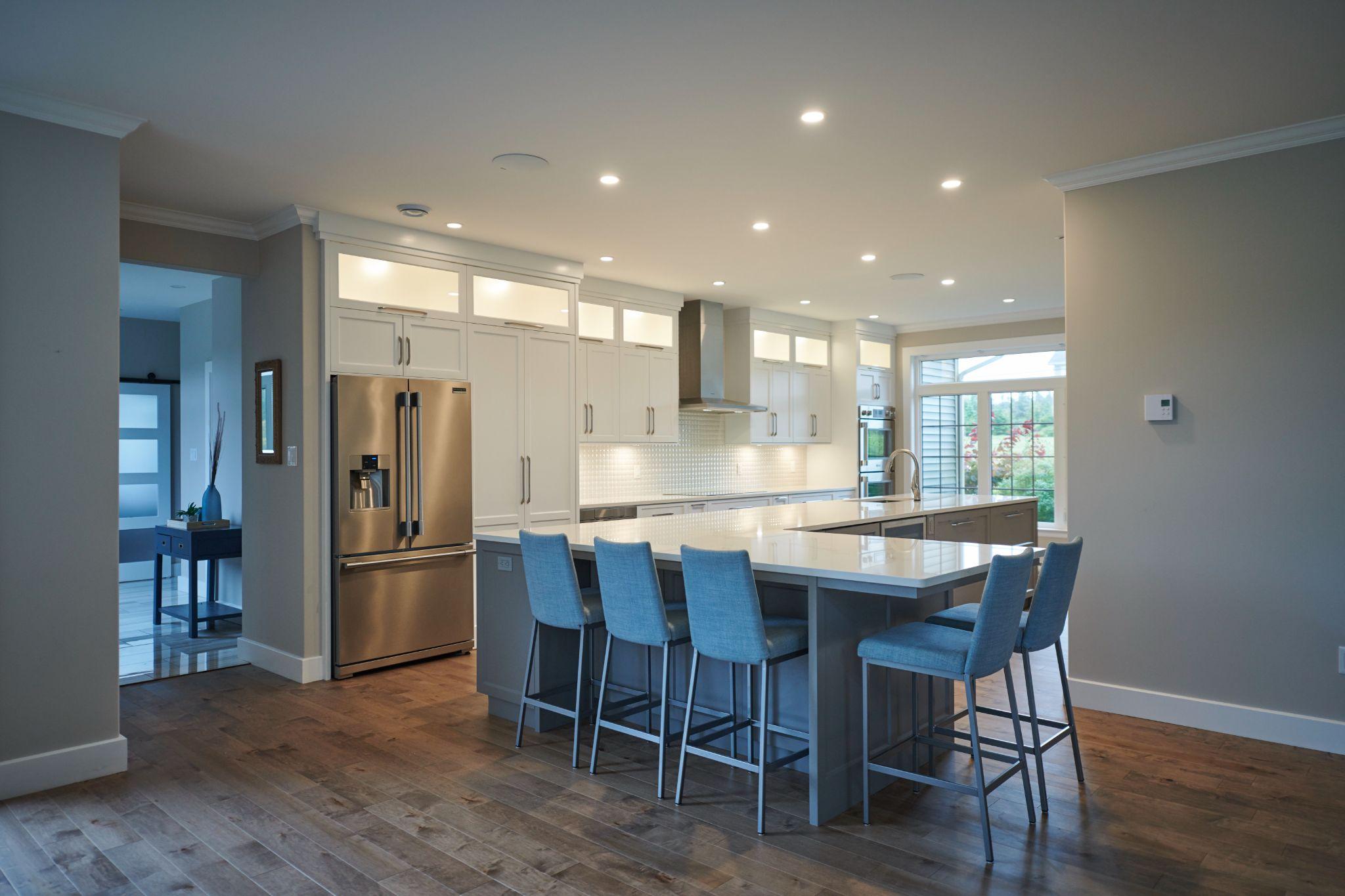 Spacious kitchen with a large island featuring blue upholstered bar stools, stainless steel fridge, and modern white cabinetry with under-cabinet lighting.